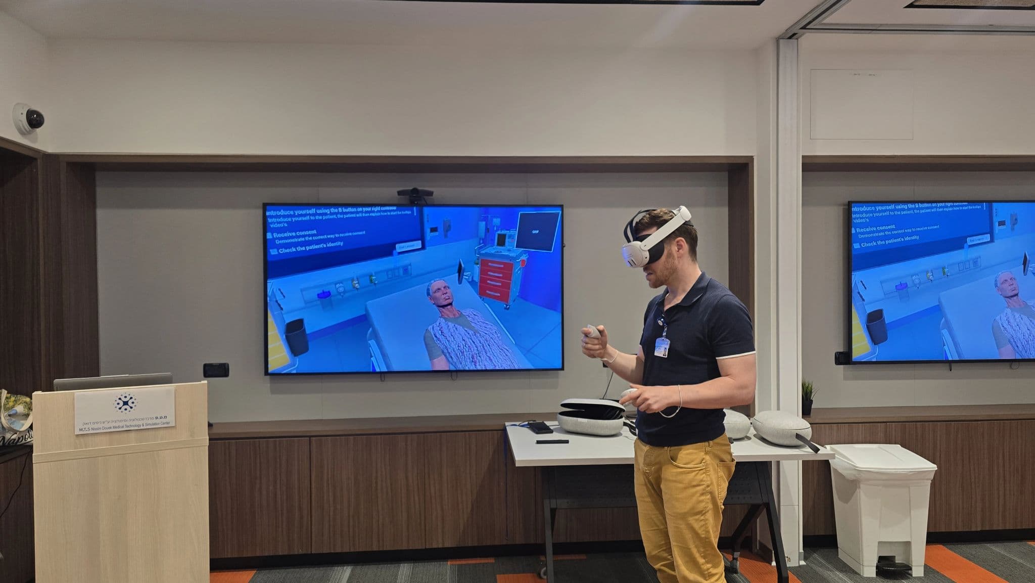 A learner in a VR headset running an InvolveXR scenario at a hospital simulation center, the scene mirrored on wall screens showing consent and patient-identity prompts