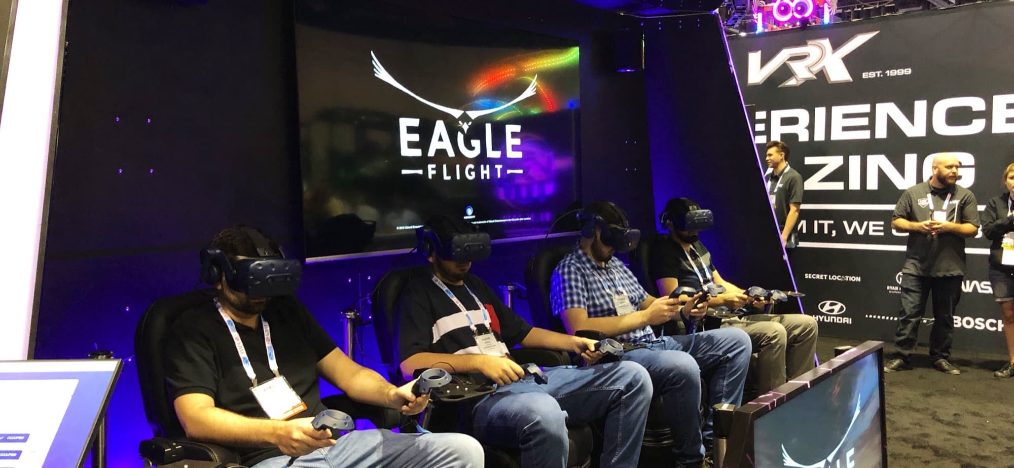 Four visitors in D-BOX motion chairs wearing VR headsets playing Eagle Flight at the IAAPA Expo booth, with the Eagle Flight logo on the big screen behind them