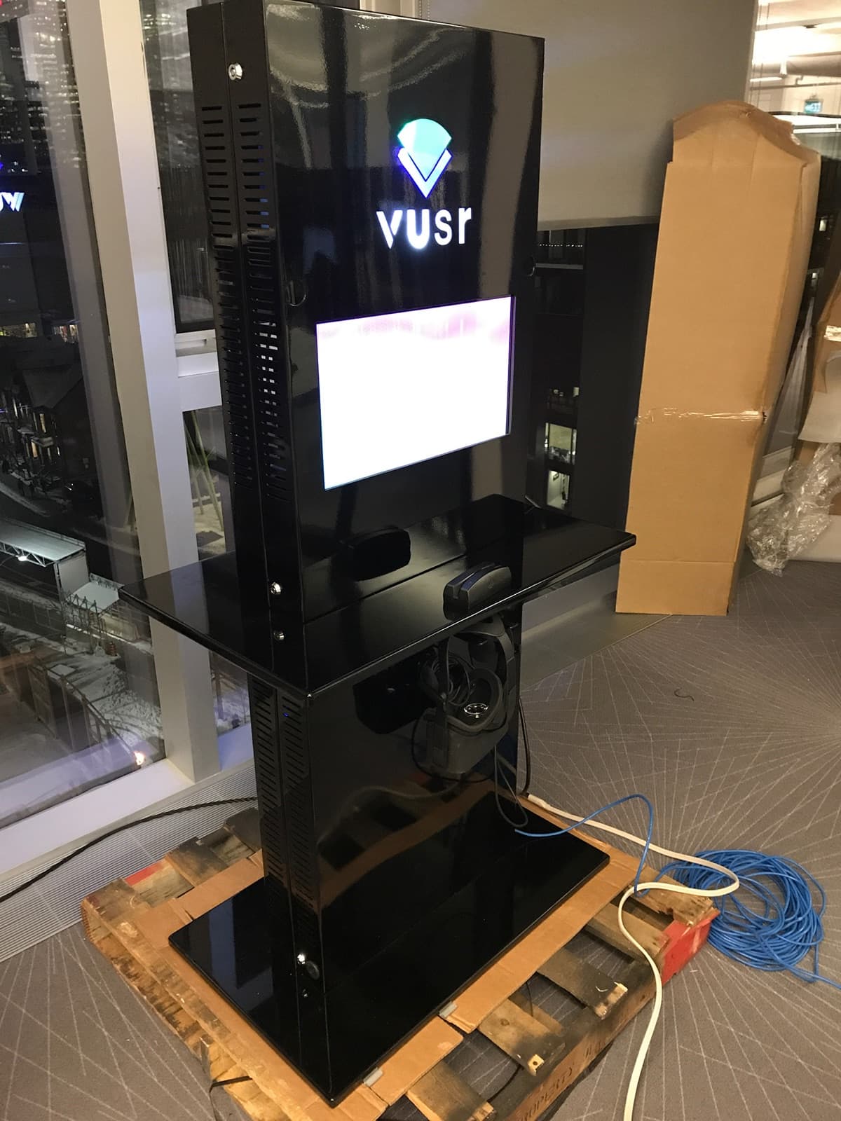 The finished Vusr kiosk on a shipping pallet: glossy black tower with illuminated Vusr logo, built-in screen, shelf with card reader, and a headset docked below